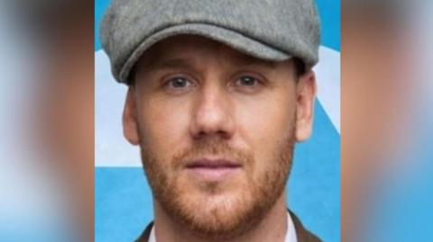 A man looking into the camera. He has a light beard and is wearing a grey flat cap. Behind him is a light blue background with white shapes on it.