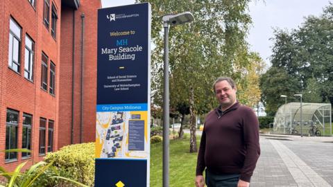 Dr Stone is standing next to a university of wolverhampton sign, which is tall and navy blue. It has a map of the campus and says "Mary Seacole building". Dr Stone has short brown and grey hair and is wearing a maroon jumper.