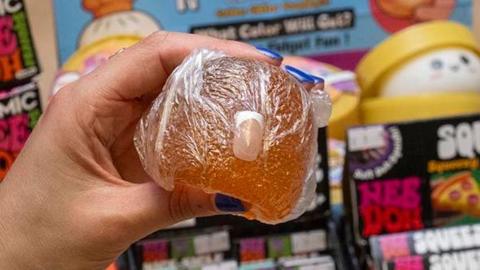 An orange squishy toy is being held, and a false fingernail is protruding from it.