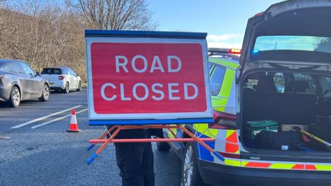 A police officer holds a red 'road closed sign' next to a police car on the A38. 