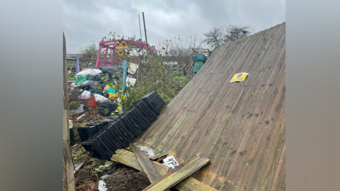 Damaged wooden fencing, which is leaning backwards.  There is lots of mud and foliage in the background.  It is also cloudy.