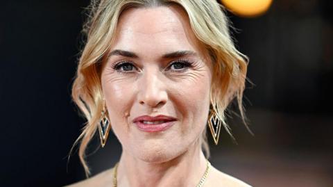 A head and shoulders image of actress Kate Winslet. She has short blonde hair and is wearing a gold necklace.