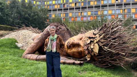A woman with short grey hair stands in front of a large wooden sculpture of a resting woman. She wears a suede jacket, colourful scarf and dark blue jeans. The wooden sculpture is part of a park installation.