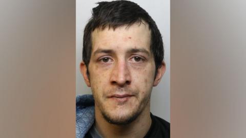 A police photograph of a man with dark hair and brown eyes. He is wearing a hooded top and looking straight at the camera. 