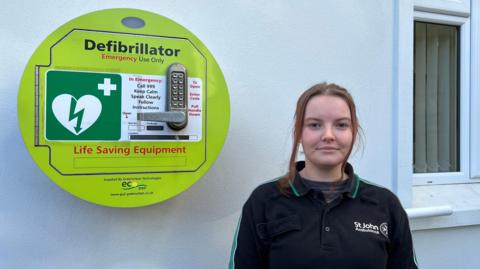 A round green wall-mounted defibrillator case labeled “Defibrillator – Emergency Use Only” with instructions to call 999 and follow directions. It features a white heart symbol with a green lightning bolt and a cross, indicating medical emergency equipment. The case has a keypad lock and a handle for access, and the words “Life Saving Equipment” are printed in red at the bottom. A woman is standing next to it wearing a St John Ambulance work shirt.