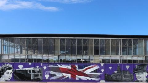 A mural depicting Queen Elizabeth II's likeness in 1952 and 2022, Great Yarmouth's Town Hall, a Union Jack symbol and a a picture of the train that brought her to Great Yarmouth in 1985. The paintings are on a purple background. 