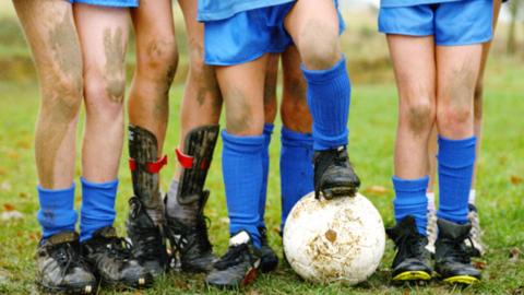 Four children stand in a line after a football match wearing blue socks and black football shoes. One of them is standing on a white soccer ball.