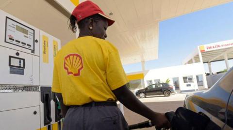The back of a woman wearing a yellow T-shirt with a Shell logo on the back and a red cap stands by a car as she fills it up with petrol.