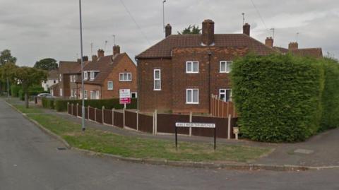 One end of a street of two-storey brick houses with white-framed windows, enclosed by low wooden fences. There is a white street sign saying "Whitworth Avenue" in the centre of the shot. There is a tall hedge to the right and a road with a grass verge sweeps round to the right.