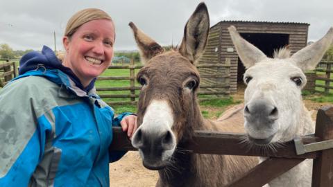 Louise Stuart has blonde hair and is wearing a blue coat. She is smiling and standing next to two donkeys 