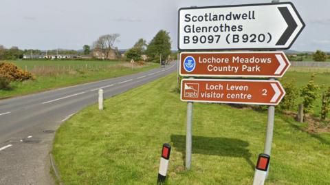 Picture of a road sign with a country road in the background. It features thee signs. One, which is white with black writing says "Scotlandwell Glenrothes B9097 (B920). Below this sign is two brown signs with white writing. One says: "Lochore Meadows Country Park" while the other says "Loch Leven visitor centre 2". All three signs are pointing the left. 