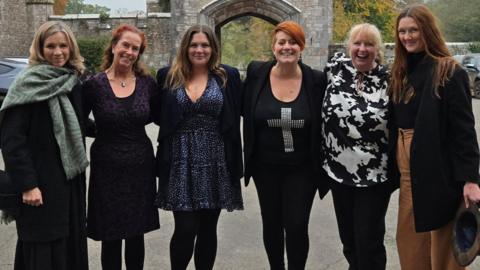 Directors of Phoenix Rising CIC. The women are standing in a line and have their arms around each other. They are standing in a car park and are smiling at the camera. Some of the ladies are wearing dresses while others are dressed in trousers and tops. 