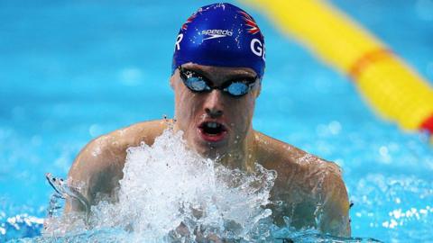 A swimmer in a UK blue Speedo hat comes out of the water in a pool.