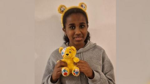 Bethel smiling holding a yellow Pudsey bear with yellow Pudsey bear headband. She is wearing grey hoodie. 