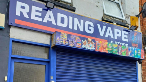 A general view of Reading Vape, which is has blue shutters, a blue door on the left of the picture and a blue sign with white lettering that spells out "Reading Vape and Store". 