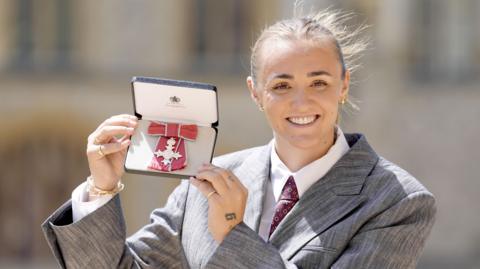 Georgia Stanway wearing a grey suit and holding up her MBE. She has a broad smile and her hair is tied back.