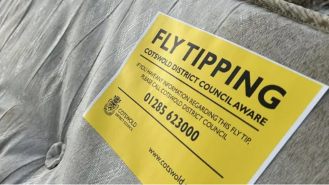 A discarded mattress with a yellow sign saying Fly tipping, Cotswold Disctrict Council aware. And it holds contact information of 01285 623000 to get in touch with the authority if you have any information regarding that waste.