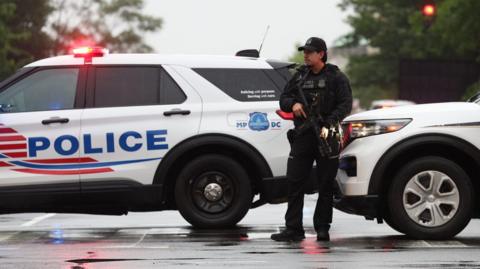A police officer with a gun stands in front of two white marked police cars