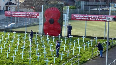 Four men standing in a grassy area where there are rows of white crosses and a banner of a large poppy with two banners on either side