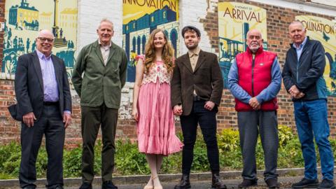 From left to right: Councillor Nigel Cooke, cabinet member for environment, leisure and culture at Stockton-on-Tees Borough Council (he is bald and wearing glasses, a blue shirt and a grey suit), Reuben Kench, chair of the Stockton & Darlington Rail Heritage Partnership (he has grey hair and is wearing a green suit jacket), Abby (she has strawberry blonde hair and is wearing a pink sundress with a knitted vest top), Owen (he has light, brown hair and is wearing a flat cap and a brown suit jacket, Councillor John Coulson (Ward Councillor for Yarm) (he is bald with a grey beard and mustache, and is wearing a blue, long-sleeved shirt and a red puffer coat)and Councillor Andrew Sherris (Ward Councillor for Yarm) (he has grey hair and is wearing a blue shirt with a grey raincoat). They are standing in front of the mural, West Street, Yarm. The mural is on the side of a brick building, and consists of eight individual panels that display words and images associated with the railway. Some of the words visible include: together, innovation, arrival, history, etc.