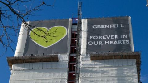 A black and white sign covers Grenfell Tower which reads 'Grenfell, Forever in Our Hearts', with a clear blue sky in the background. 