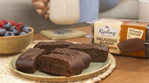 A plate with five Mr Kipling chocolate cake slices on, with the packet behind it. There is also a bowl of berries on the table next to the cake.