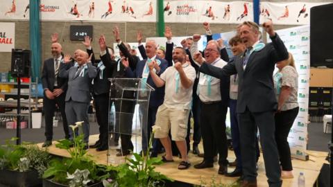 Lots of men in suits with blue rosettes, and one woman, stand on a stage cheering with their arms raised in triumph. 