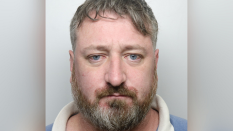 Custody picture of a man with blue eyes and a full beard who looks directly at the camera. He has short, slightly tousled hair and wears a collared shirt, with a plain light background behind him.