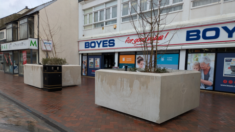 Two large stone squared plant pots on a high street. Small leafless trees grow from them. They are outside a Boyes shop and it has been raining.