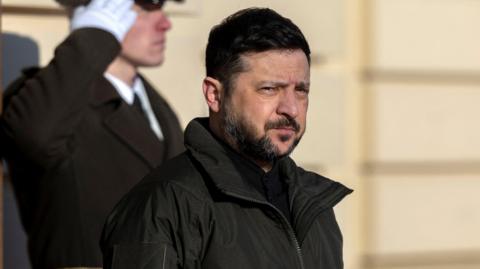 Zelensky, who has short black hair and short black facial hair, wears a black coat in front of a building with beige walls. Behind him a man in uniform stands to attentoin.