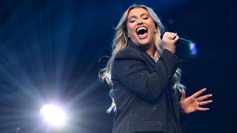 Ella Henderson is on a stage with a spotlight shining behind her. She has long blonde hair and is wearing a sparkly suit jacket as she sings into a microphone.