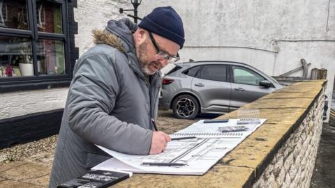 A man is wearing glasses, a grey thick coat and a navy beanie, and he has a sketchbook open in front of him, on a wall. He is drawing a building in pencil. A window of a building is behind him and there is a white wall and a grey car in the background.