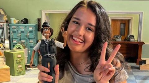 Lauren Patel holding a figurine of PC Mukherjee, smiling in a studio. She is wearing a grey top and has long brown hair. 