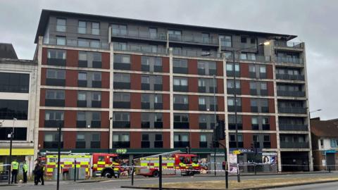 The red and grey apartment block has seven floors, with two fire trucks outside and a police car. The seventh and sixth floor been burnt and whole building evacuated.