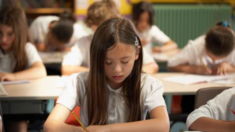 A young girl with long-brown hair holds a yellow and black pencil in her hand. She wears a white polo top. Behind her are five young people in the same outfit, holding similar pencils writing on white pages. 