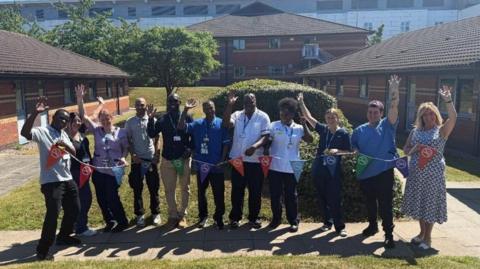 A group of 11 NHS staff stand outside and wave at the camera while holding multi-coloured bunting, which has stop smoking symbols on it.