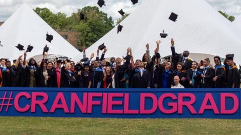 A large group of students dressed in graduation cloaks throw their hats into the air. They are outside on a grassy area with two white marquees behind. In the foreground is a large sign in pink reading #CranfieldGrad, against a blue background.