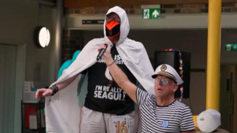 A man dressed as a seagull with a white cloak and a orange beak. Another man in a striped top and a sailor hat is holding up a microphone to the man's face.