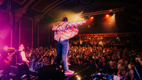 Richard Reeves is seen standing on a piano on a stage facing a crowd of people.