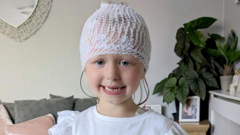 Noa-Rose smiling looking at the camera. She wears a white t-shirt and has a bandage around her head with colourful wires coming out at the back. 