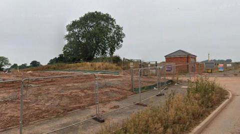 A housing development site with brown mud in an open field, fenced off by metal fencing. There is a small brick structure and a road running through the site