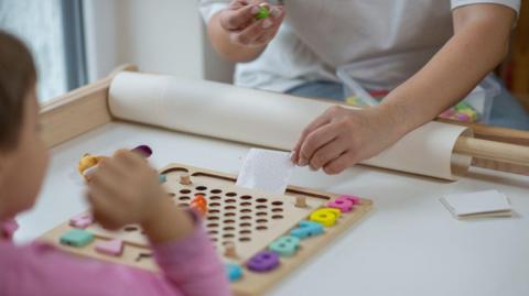 A stock photo of a blurred child in a long pink top who can be seen holding up their hand and looking at wooden boards with holes in it and colourful numbers down the side and shapes. An older person is holding a paper with circles on it at the front of the wooden board and is also holding something green and has a plastic box rested in their lap.