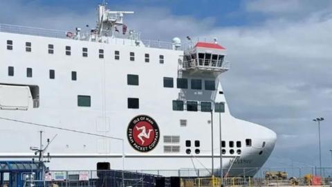 The bow of the Manxman, which is a tall white angular ferry, with the Isle of Man Steam Packet's logo on the side, which is the three legs of Man with the words Isle of Man Steam Packet Company around it.