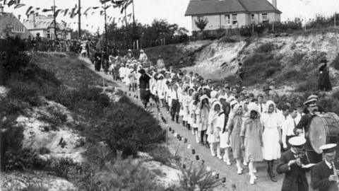 Black and white photo of a procession of children being led through a park by a marching band