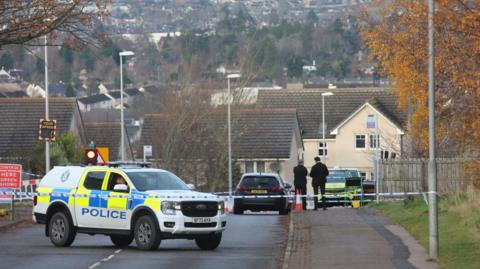 A police four-wheel drive vehicle is parked across a road. There is also blue and white police tape across the road just in front of the vehicle. Further down the road two men in dark suits stand next to each in conversation. There is a black car parked next to them and a police car in front of them. The picture has been taken looking down a hill and there are rows of houses to the bottom of the hill.
