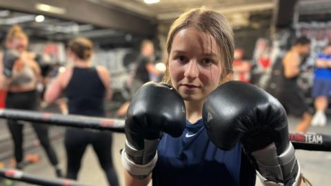 A 19 year-old woman is standing next to a boxing ring. She is wearing black boxing gloves which she is holding up close to her face. She has light brown hair tied back and is wearing a navy T-shirt. Other poeple are training in the ring behind her.