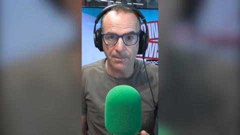 Martin Lewis is wearing dark green T-shirt and black-framed glasses. He is wearing headphones and sitting behind a green mic.