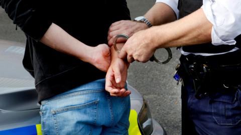 A stock image showing a police officer putting handcuffs on a man as he leans over the hood of a police car. The man is wearing blue jeans and a black hoodie and has his hands behind his back.