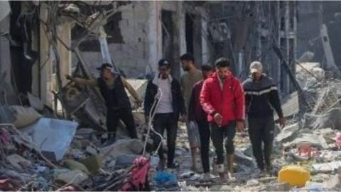 A group of people walking through rubble, outside destroyed buildings in Khan Younis on 7 April