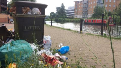 Overflowing litter bin by a canal in Northampton with bags of rubbish surrounding it.